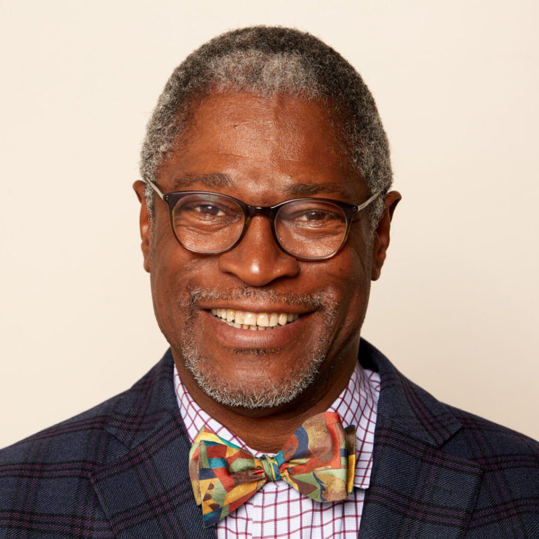 A man with glasses and bow tie in front of a white wall.